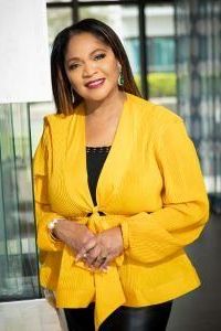 Pamela McCauley in a yellow shirt, hands folded, standing in a modern office environment.